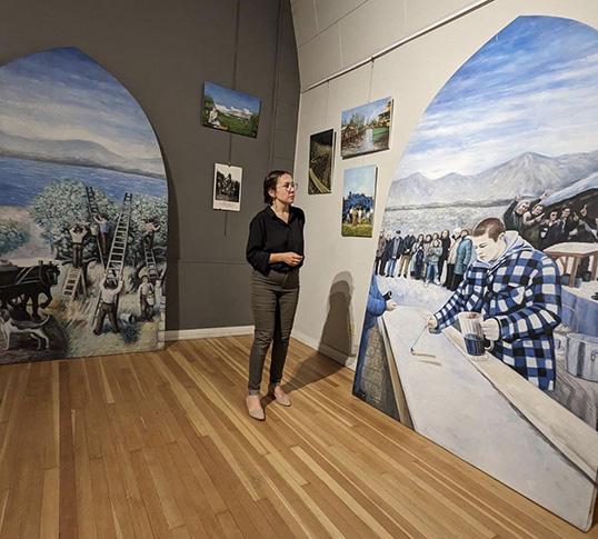 Mme Hélène Girardot, directrice du Centre Culturel Francophone de l'Okanagan, devant des toiles historiques qui racontent l'arrivée et l'établissement des francophones dans la région. (Crédit : CCFO)  